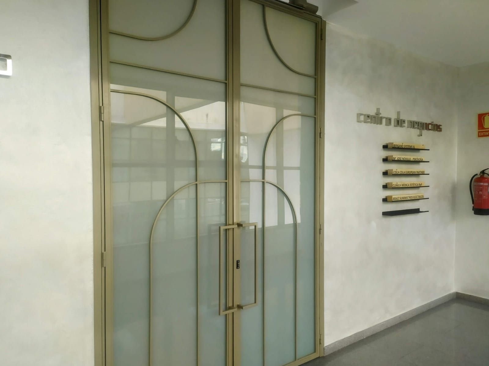 Puertas dobles de vidrio con marco y tiradores metálicos. A la derecha hay un extintor y una decoración de pared.
